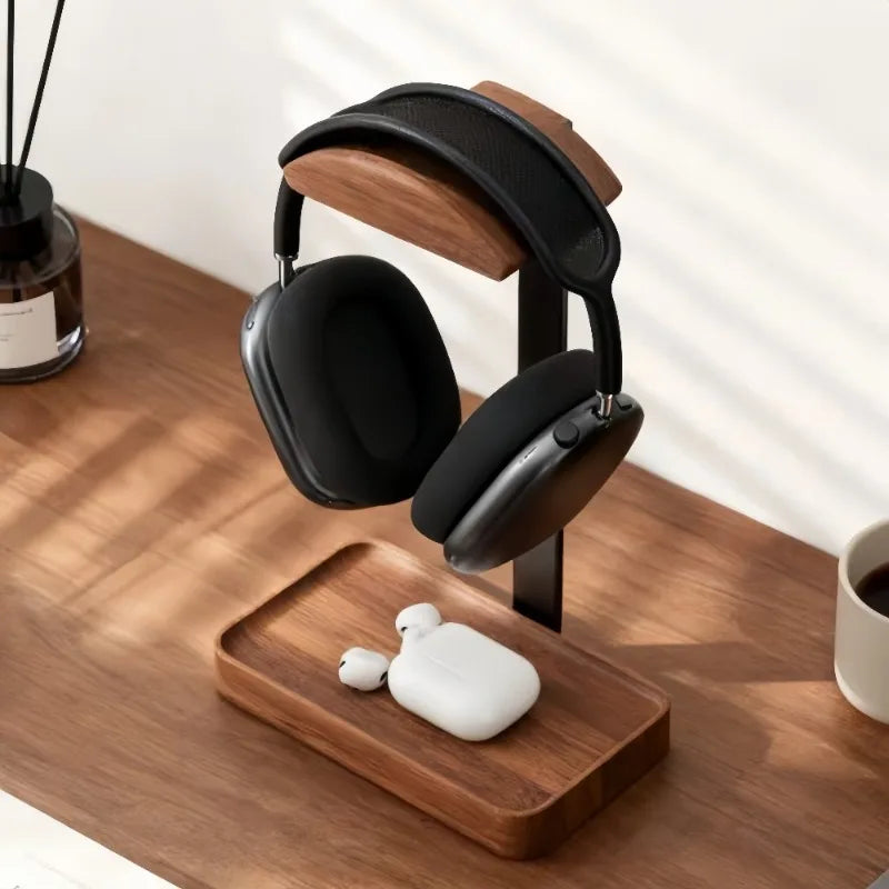Black over-ear headphones displayed on a wooden stand on a wooden table, with a white wireless earbud case and earbuds resting on the stand below. The scene is styled in a bright room with soft window shadows.